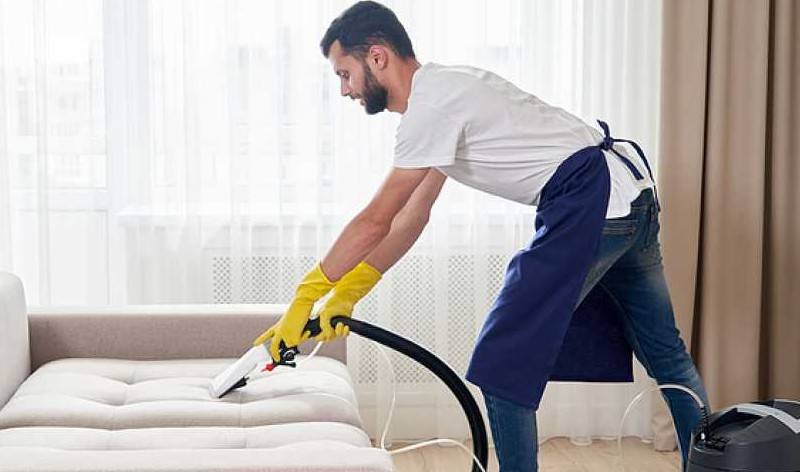 Man in white t-shirt, blue jeans, yellow gloves vacuuming sofa inside a room