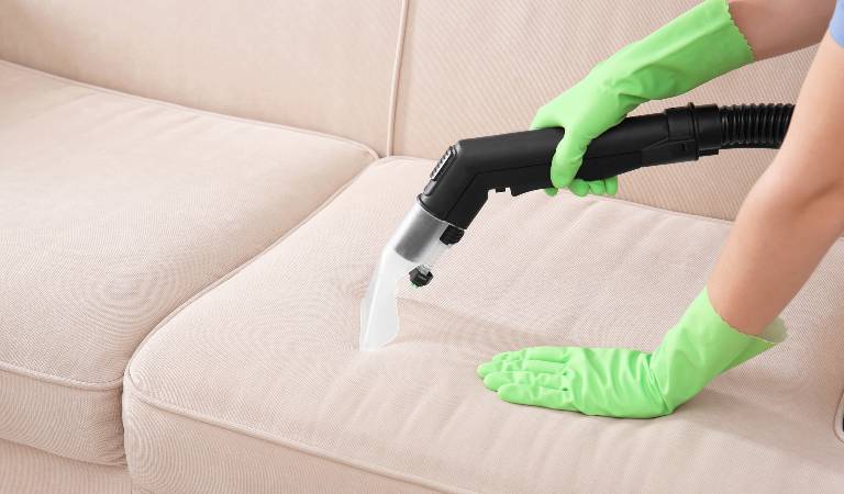 Hand of a woman in green gloves using vacuum cleaning on upholstery sofa.