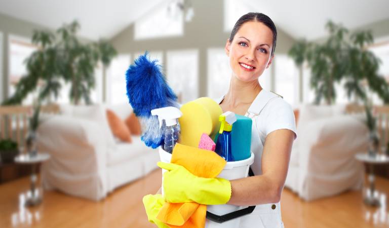 Woman in white uniform and yellow gloves holding a white basket filled with cleaning products.