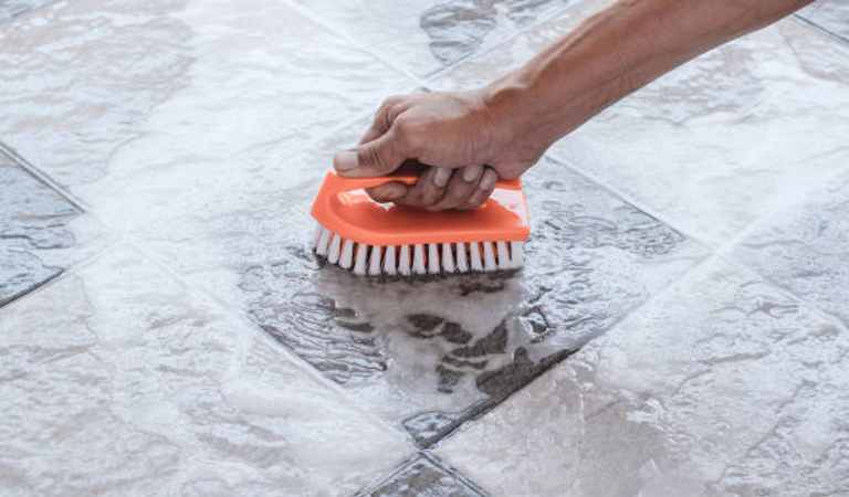 Hand of a man cleaning grout of tiles flooring using a orange brush.