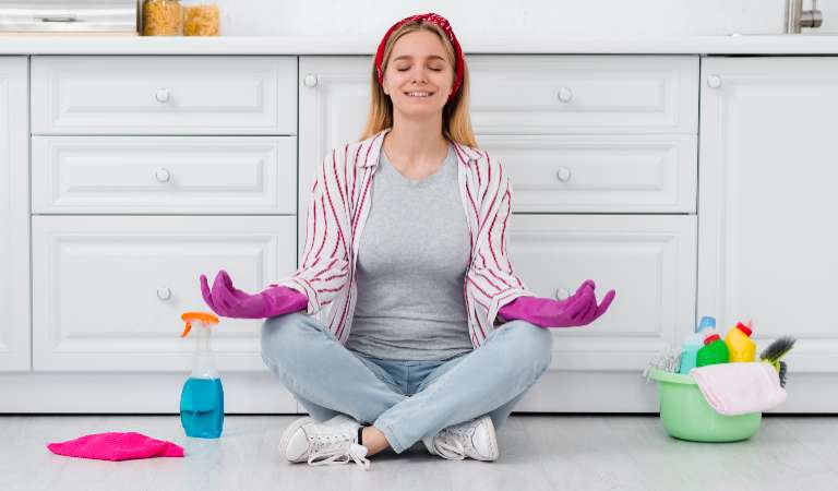 Woman in check shirt and pink glove doing yoga with a green cleaning basket and a bottle on the floor.
