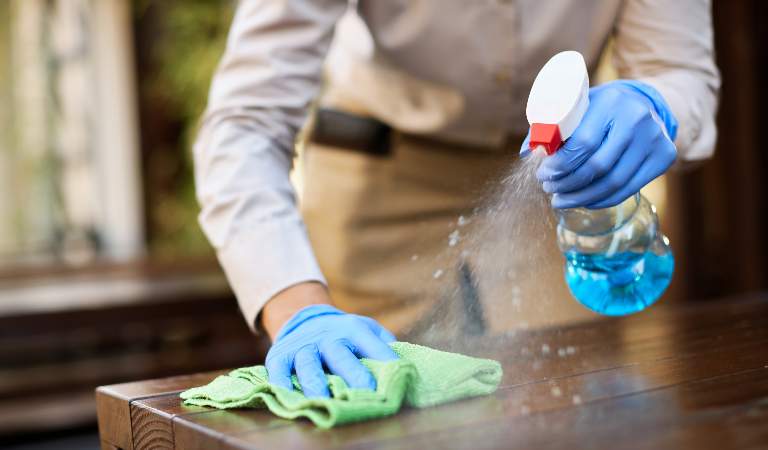 Woman cleaner in blue glove holding a bottle and cleaning table with a green cloth