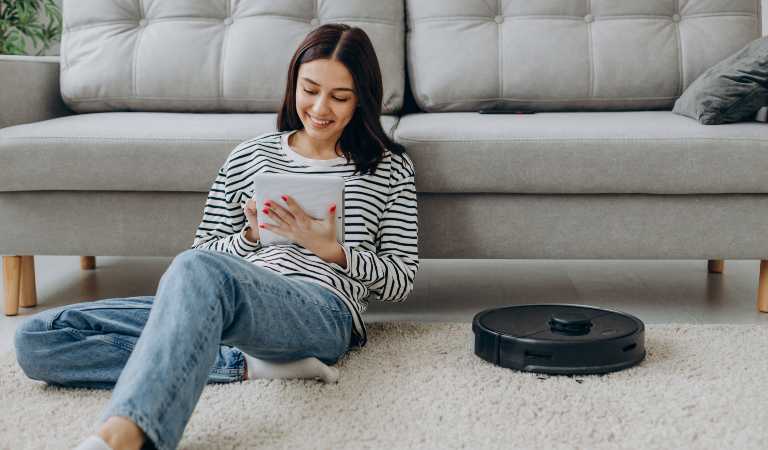 Woman is using tablet on floor with a robot vacuum cleaner on the carpet.