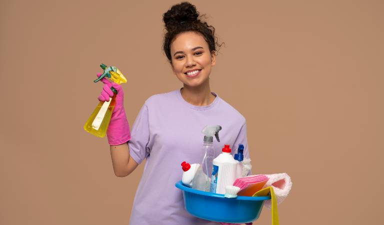 Woman in light purple top and pink gloves holding a bottle in her hand and a blue basket in her second hand.