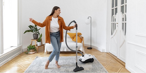 Woman in maroon long jacket and blue jeans holding a vacuum cleaner in her hand