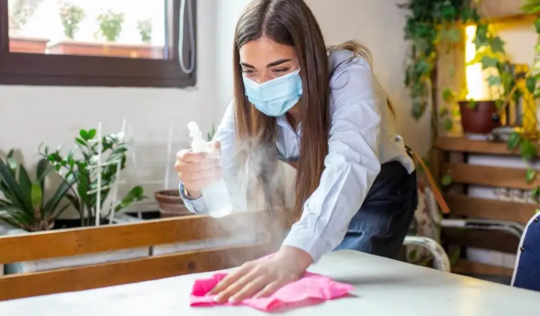 Lady in mask cleaning a table with a pink cloth holding a bottle in her hand.