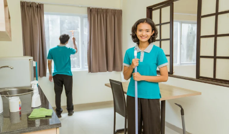Asian female janitor wearing turquoise uniform standing holding broom in room