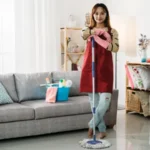 Full length asian female housekeeper standing with leg crossed is smiling at camera with confidence while keeping her arms on the mop in a bright home interior