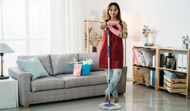 Full length asian female housekeeper standing with leg crossed is smiling at camera with confidence while keeping her arms on the mop in a bright home interior