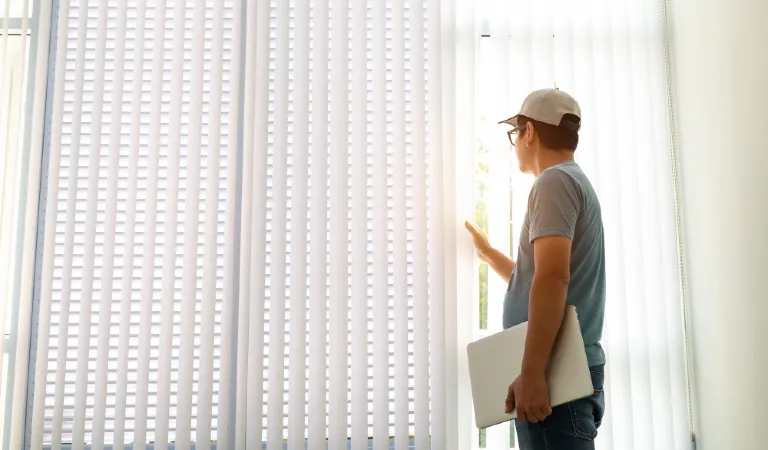 Man looking at the vertical window blind and holding a laptop in his hand