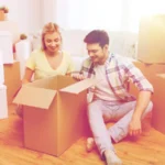 Moving, people, repair and real estate concept - smiling couple looking into big cardboard box at new home